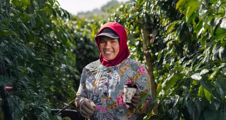 Petani kopi tersenyum memegang gelas Kopi Kenangan di antara tanaman kopi